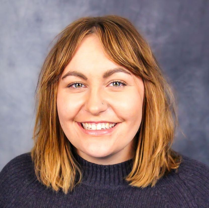 Image of a smiling caucasian woman wearing a purple sweater. She has shoulder length brown hair and is standing in front of a grep backdrop.