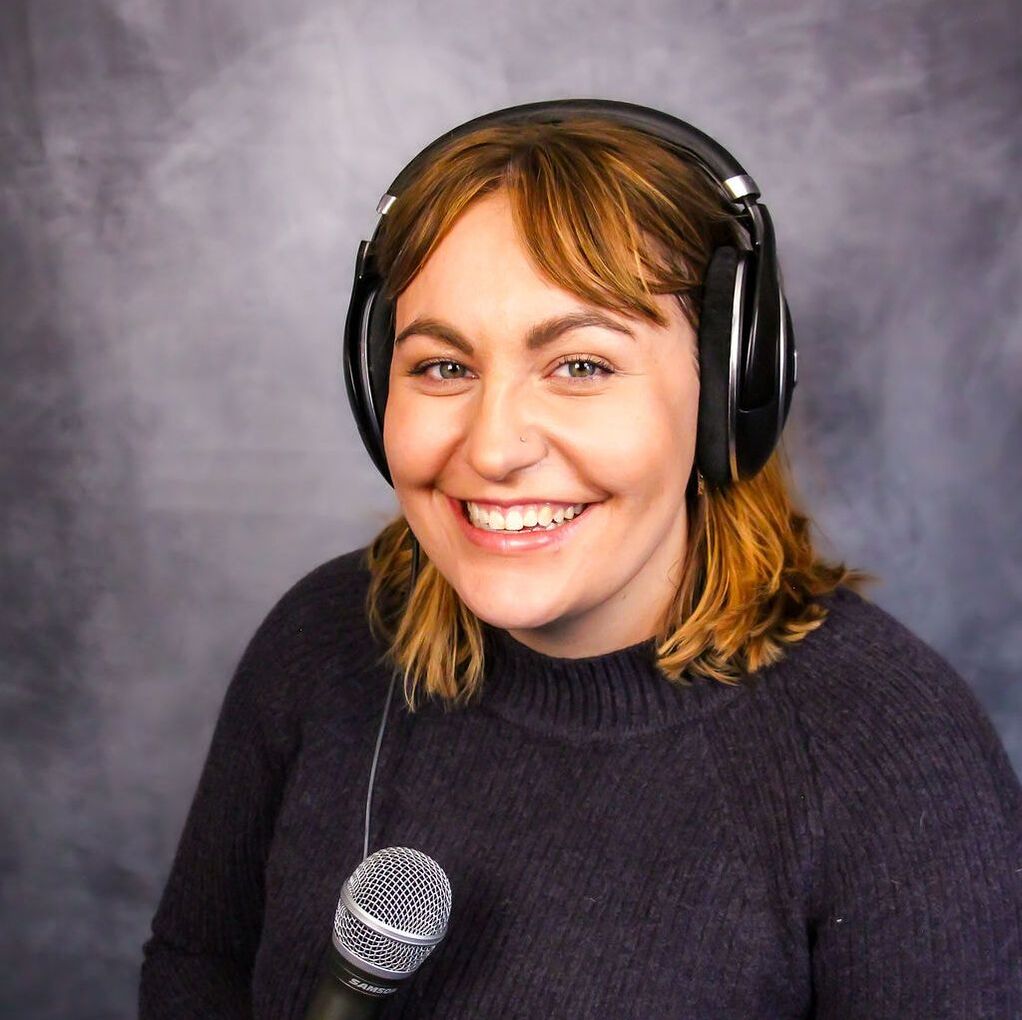 Image of a smiling caucasian woman wearing over-ear headphones and a purple sweater in front of a grep backdrop. She has should-length brown hair and is holding a microphone.