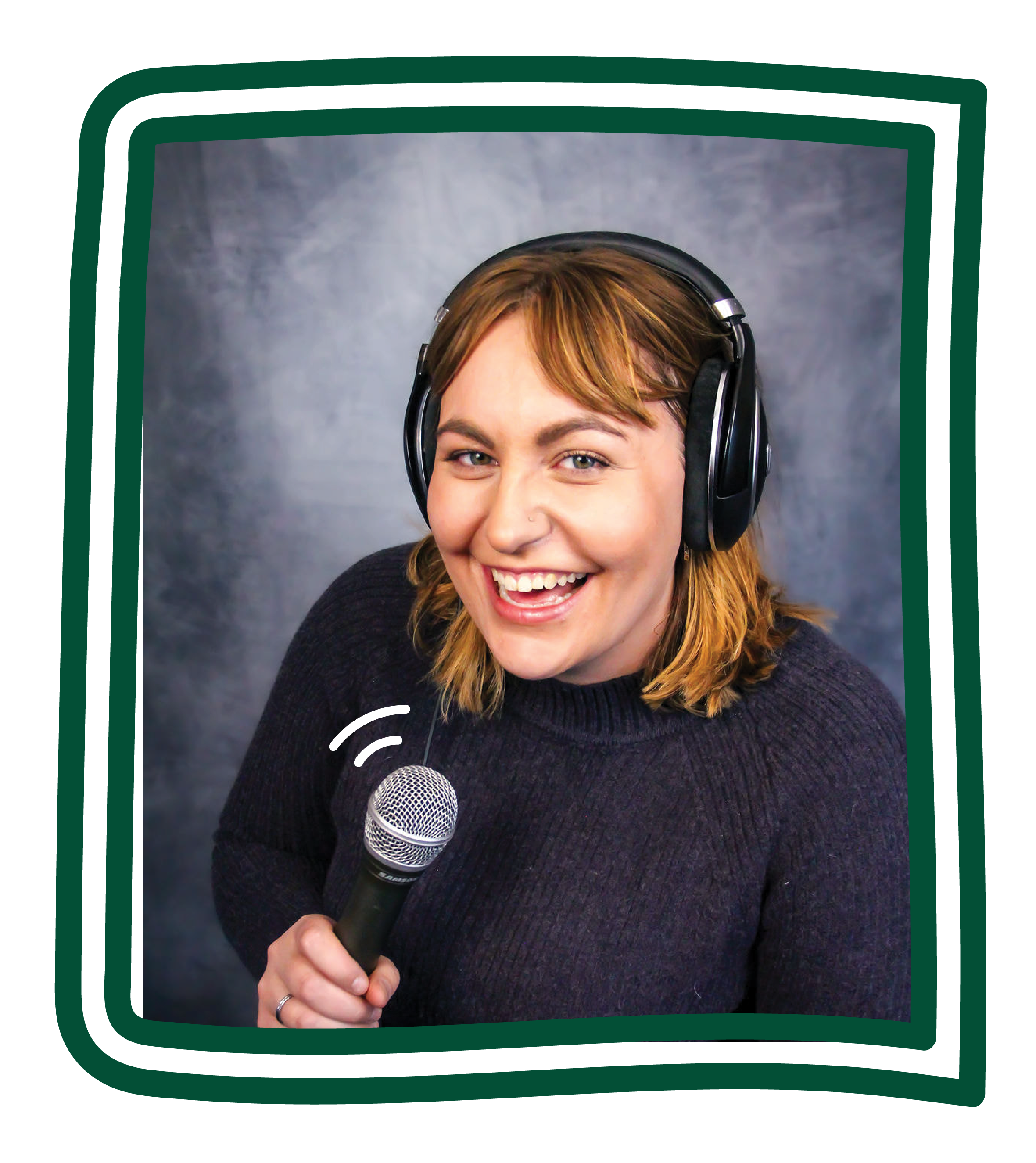 Image of a smiling caucasian woman wearing a purple sweater and holding a microphone. She has shoulder length brown hair and is standing in front of a grep backdrop.