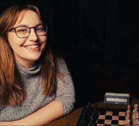Image of a smiling woman wearing a grey turtleneck long-sleeved shirt. She has long brown hair and is seated in front of a chess board, pictured to the right of the foreground.